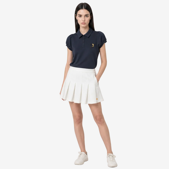A woman modeling a navy blue polo shirt with a small gold embroidered logo on the chest, paired with a white pleated tennis skirt and white sneakers, against a light gray studio background.