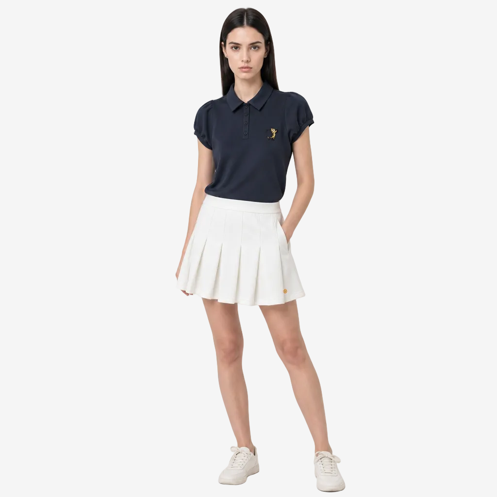 A woman modeling a navy blue polo shirt with a small gold embroidered logo on the chest, paired with a white pleated tennis skirt and white sneakers, against a light gray studio background.