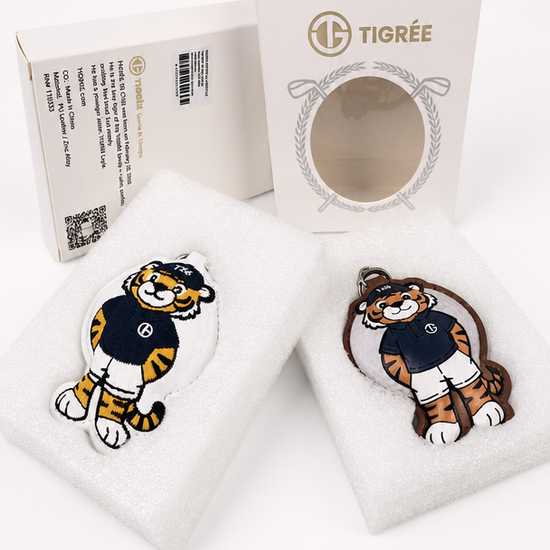 Two tiger mascot keychains displayed on white foam inserts in front of their branded packaging. The keychains feature a cartoon tiger in a navy and gold uniform, and the packaging displays the brand name 'TIGRÉE'.
