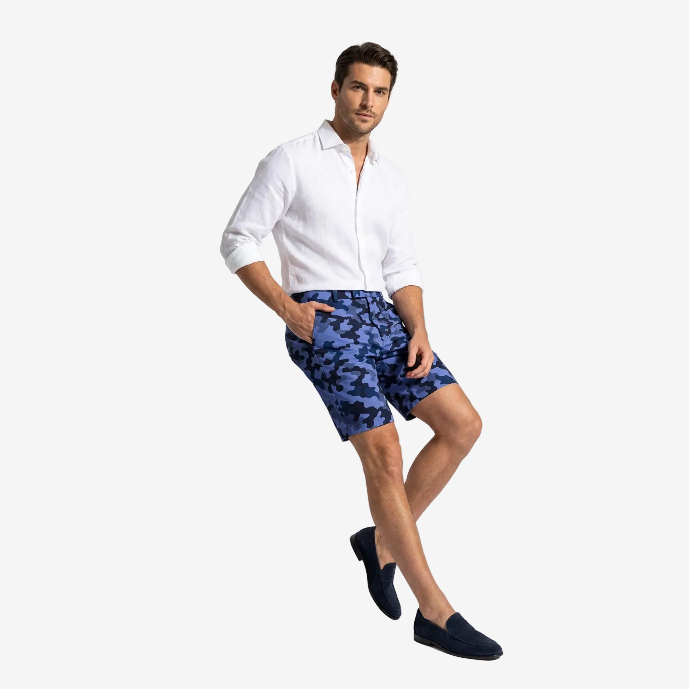 A man models a pair of blue and navy camouflage shorts, paired with a white linen shirt and dark loafers, leaning against a white block in a studio setting.