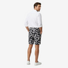 A man wearing black and white camouflage shorts, a white button-down shirt, and white sneakers, shown from a rear three-quarter view against a light gray studio background.