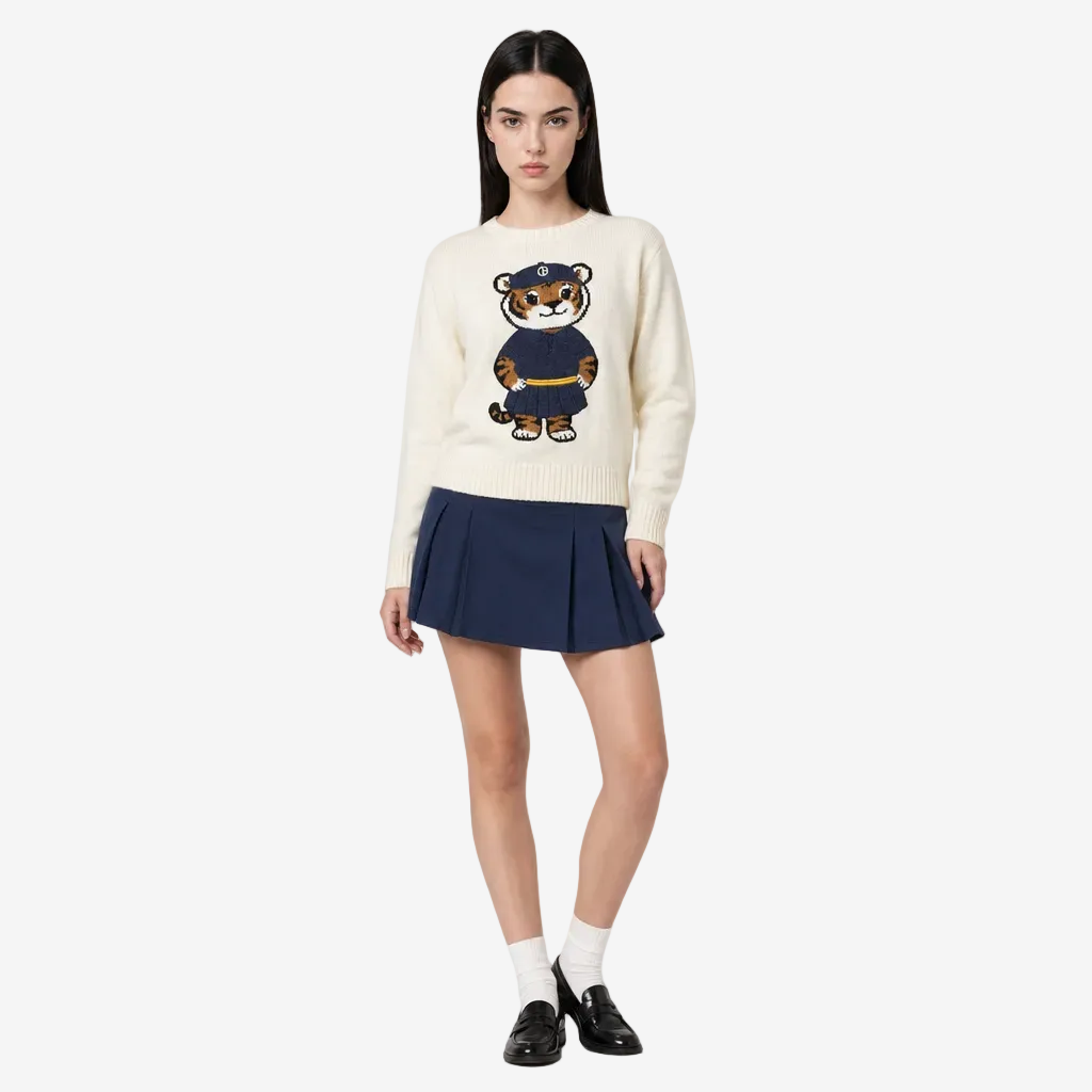 Woman wearing a cream sweater with a tiger design and a navy pleated skirt on a white background