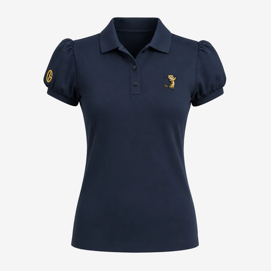 A woman modeling a navy blue short-sleeve polo shirt with a small gold embroidered logo on the chest, paired with white cropped pants and white heels, against a light gray studio background.