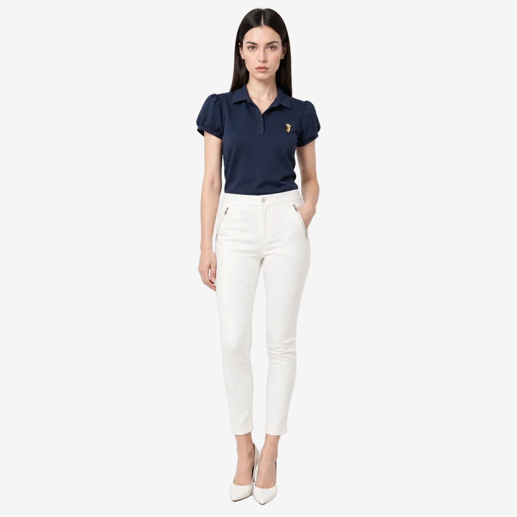 A woman modeling a navy blue short-sleeve polo shirt with a small gold embroidered logo on the chest, paired with white cropped pants and white heels, against a light gray studio background.