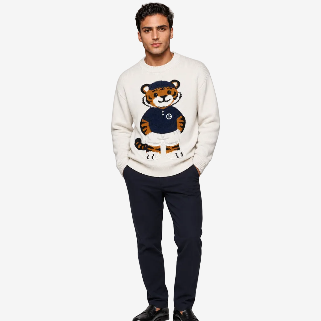 A man modeling a cream-colored crewneck sweater featuring a large embroidered tiger graphic on the front, styled with dark navy trousers and black shoes against a light gray studio background.