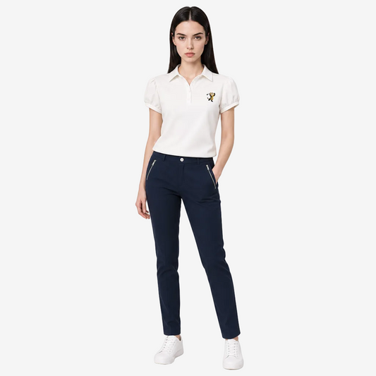 A woman modeling a white short-sleeve polo shirt with a small embroidered logo on the left chest, paired with dark navy trousers and white sneakers, against a light gray studio background.