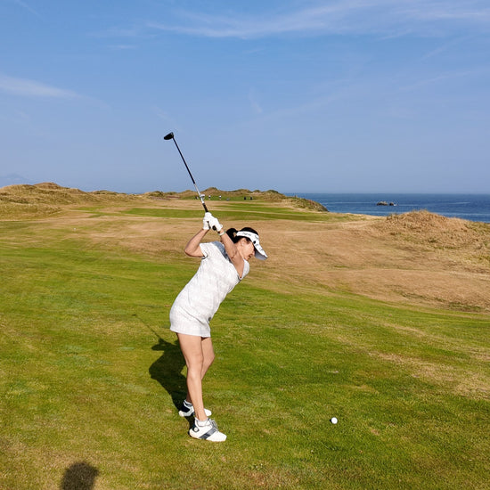 A woman in a white outfit swings a golf club on a grassy course under a clear blue sky with the ocean visible in the distance.