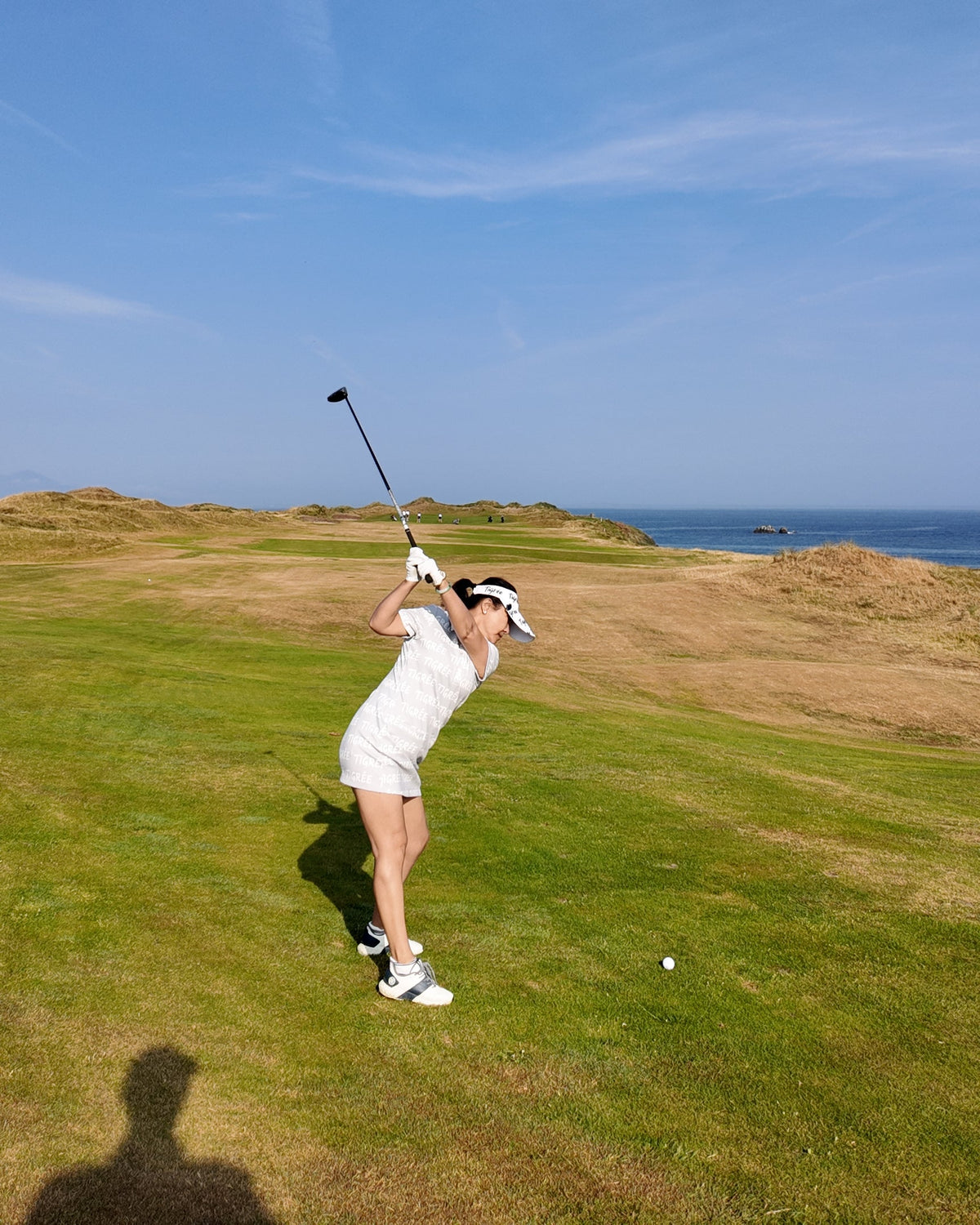 A woman in a white outfit swings a golf club on a grassy course under a clear blue sky with the ocean visible in the distance.