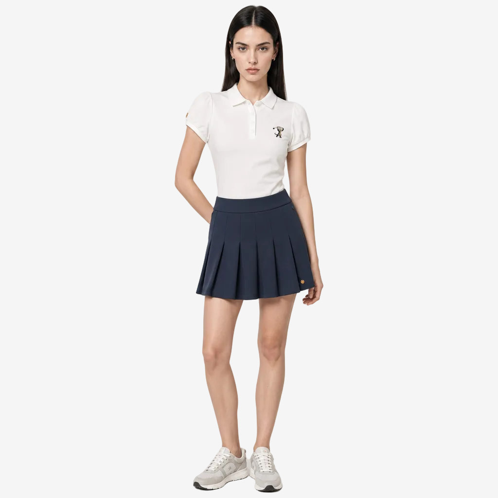 A woman modeling a white short-sleeve polo shirt and a navy blue pleated tennis skirt, paired with white sneakers, against a light gray studio background.