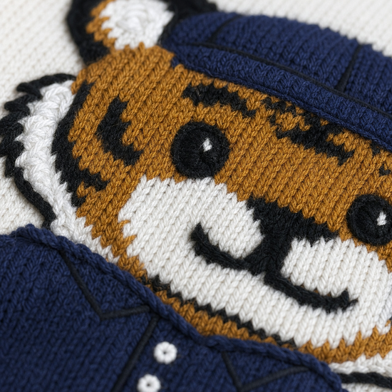 Close-up of a knitted tiger face patch or applique on a navy blue and white garment, showing the tiger's face, stripes, and a navy hat.