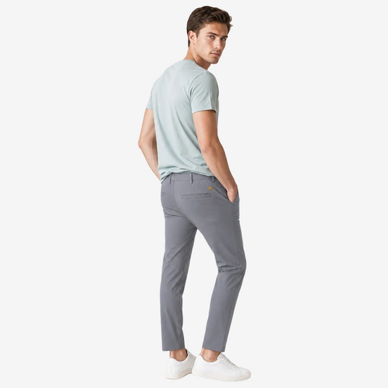 A man wearing a light sage green t-shirt and gray slim-fit trousers, standing in profile against a light gray studio background.