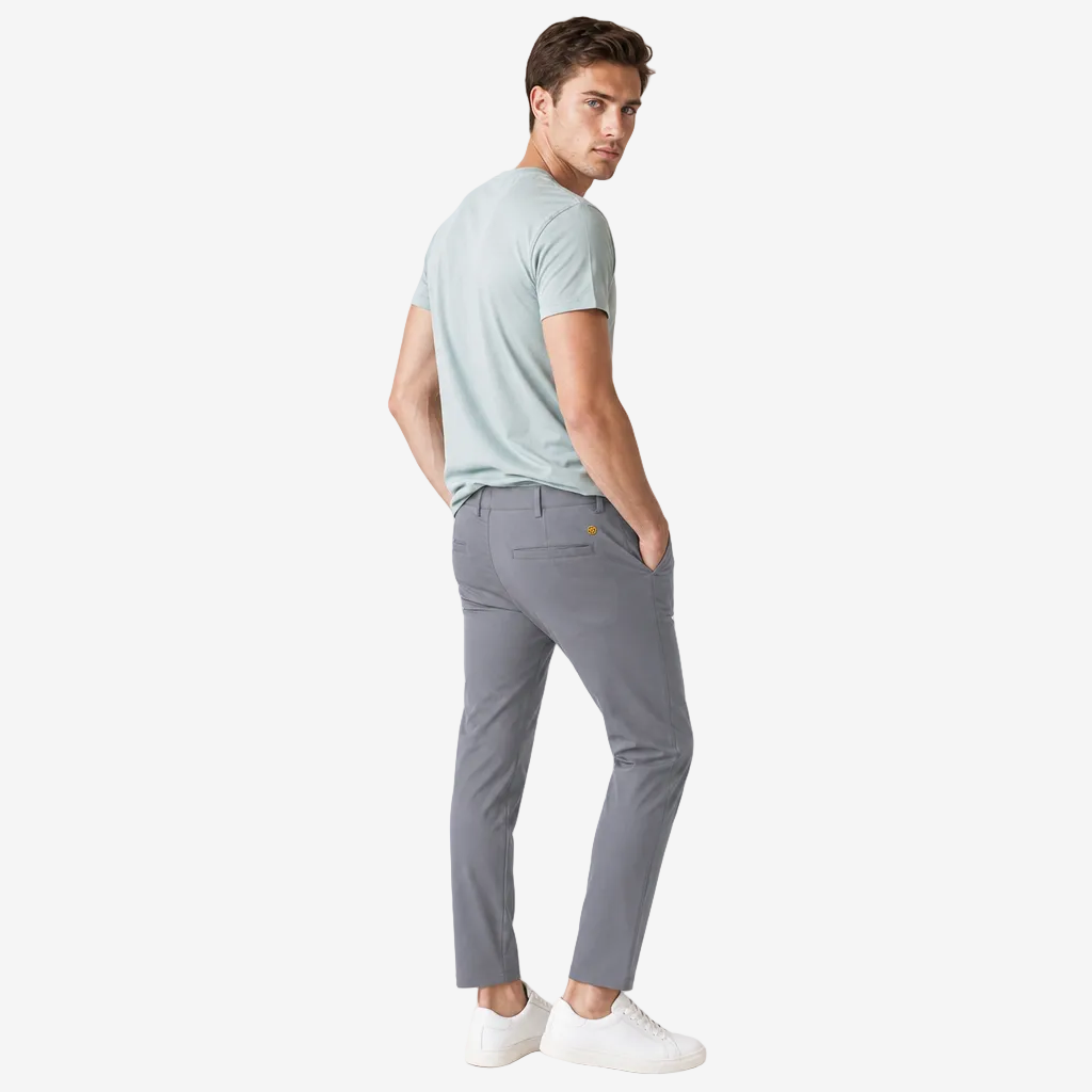 A man wearing a light sage green t-shirt and gray slim-fit trousers, standing in profile against a light gray studio background.