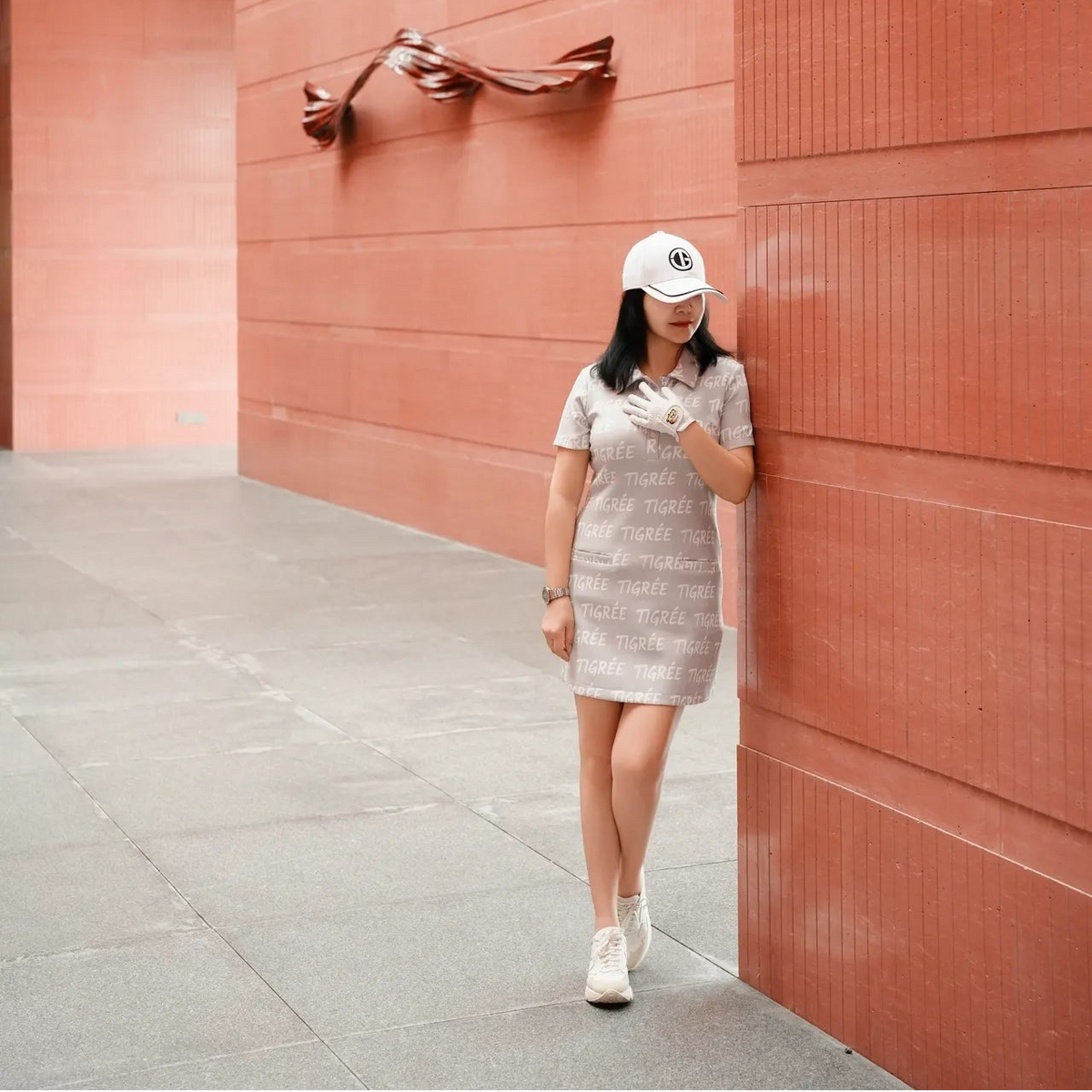 Woman in a dress and cap standing against a pink wall with a sculpture.